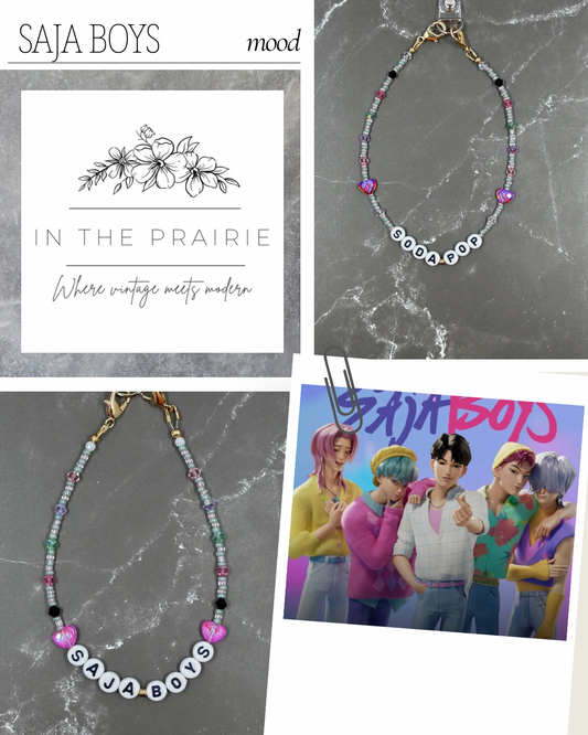 Jewelry with 'Saja Boys' branding on a gray marble background, featuring a necklace and bracelet.